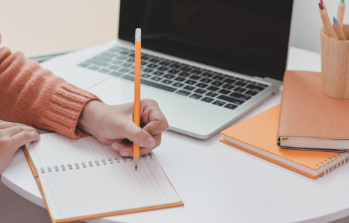 Person writing in a notebook on a desk with laptop and stationery